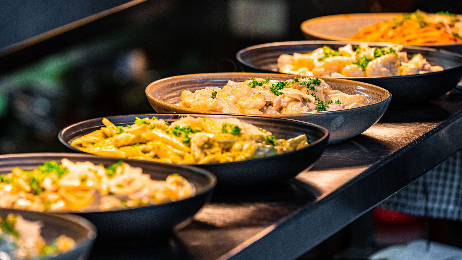 Image is of a row of black bowls filled with pasta and gnocchi under lights