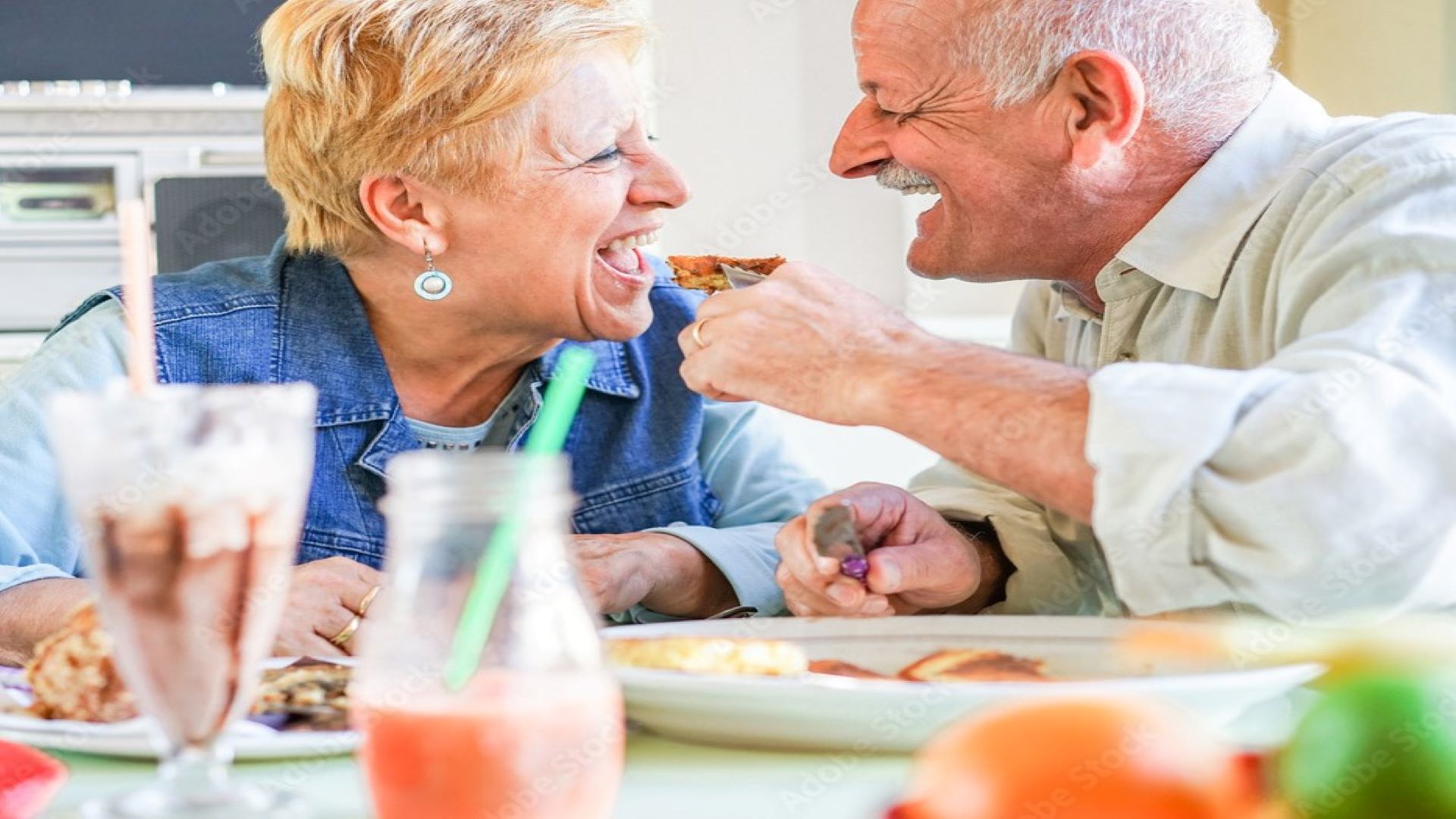 Image is of an older couple laughing and facing each other as they eat food