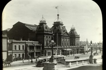 An old sepia photograph of The Princess Theatre, taken during the late Victorian era. The theatre is large and opulent, with an extravagant French-style facade of domed roofs and stained-glass windows. However, as the photograph is devoid of colours, the theatre appears black and grim, creating a foreboding presence for the otherwise beautiful building.