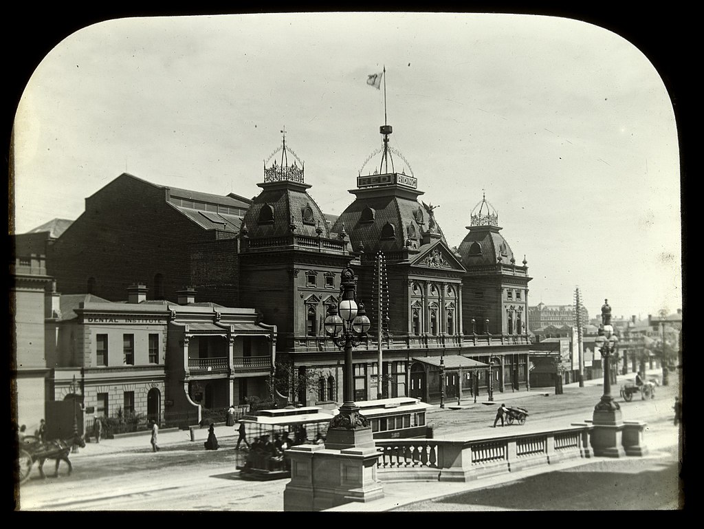 An old sepia photograph of The Princess Theatre, taken during the late Victorian era. The theatre is large and opulent, with an extravagant French-style facade of domed roofs and stained-glass windows. However, as the photograph is devoid of colours, the theatre appears black and grim, creating a foreboding presence for the otherwise beautiful building.